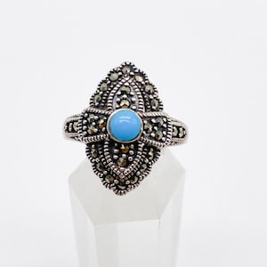 Puede incluir: Un anillo de plata con una piedra preciosa azul rodeada de piedras de marcasita. El anillo está en un expositor blanco.