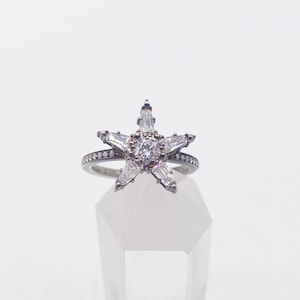 May include: A silver ring with a clear crystal star design. The ring is displayed on a clear crystal stand.