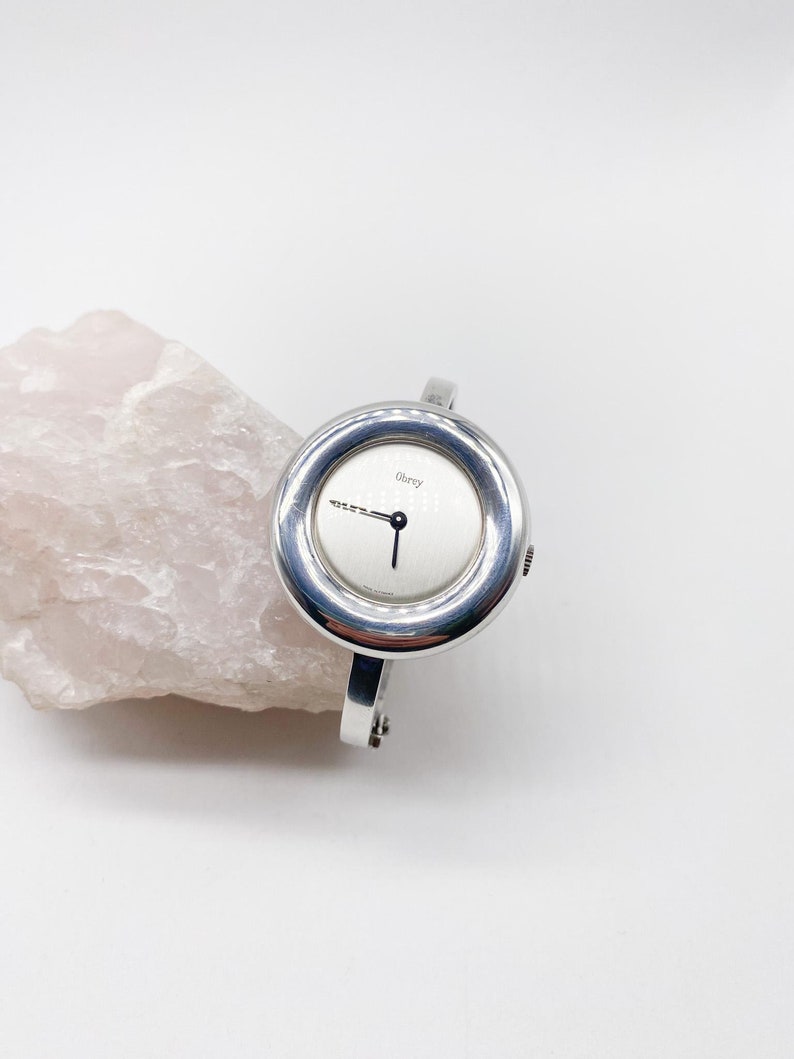 May include: A silver wristwatch with a round face and a white dial. The watch has a silver band and the word "Obrey" printed on the face.