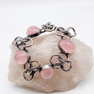 Puede incluir: Una pulsera plateada con gemas ovaladas de color rosa p&aacute;lido. Los eslabones de la pulsera tienen un dise&ntilde;o &uacute;nico e entrelazado. Las gemas est&aacute;n engastadas en un bisel. La pulsera se exhibe sobre una superficie clara y texturizada.