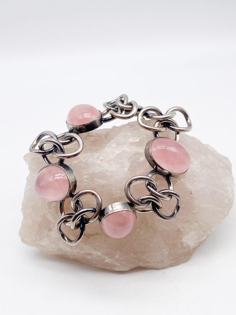 Puede incluir: Una pulsera plateada con gemas ovaladas de color rosa p&aacute;lido. Los eslabones de la pulsera tienen un dise&ntilde;o &uacute;nico e entrelazado. Las gemas est&aacute;n engastadas en un bisel. La pulsera se exhibe sobre una superficie clara y texturizada.