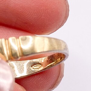 May include: Close-up of a gold ring with a textured band and a hallmark stamp. The ring has a polished finish and is held between fingers against a white background. The ring's band features vertical grooves.