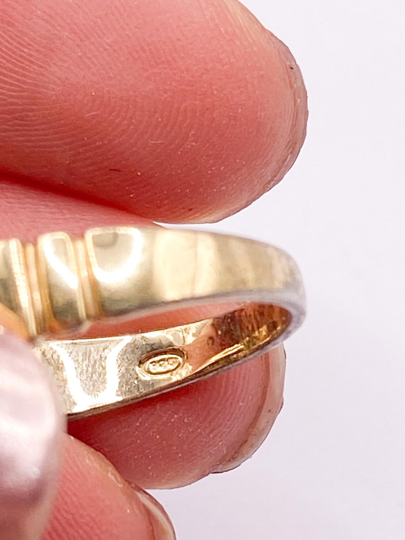 May include: Close-up of a gold ring with a textured band and a hallmark stamp. The ring has a polished finish and is held between fingers against a white background. The ring's band features vertical grooves.