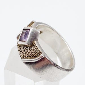 May include: A silver ring with a square, purple gemstone. The ring has a textured gold-coloured accent on the side of the gemstone setting. The band is smooth and polished. The ring is displayed on a clear, angled surface.
