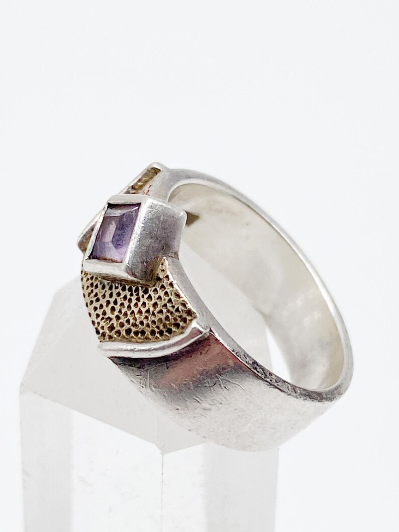 May include: A silver ring with a square, purple gemstone. The ring has a textured gold-coloured accent on the side of the gemstone setting. The band is smooth and polished. The ring is displayed on a clear, angled surface.
