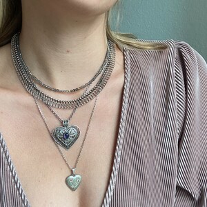 May include: A silver necklace with multiple chains and two heart pendants. The larger pendant has a blue gemstone in the center. The smaller pendant has a floral design with red gemstones.