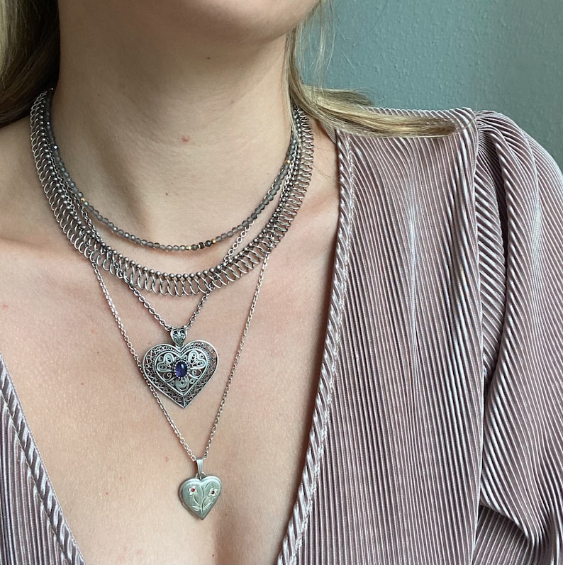May include: A silver necklace with multiple chains and two heart pendants. The larger pendant has a blue gemstone in the center. The smaller pendant has a floral design with red gemstones.