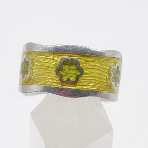 Puede incluir: Anillo de plata con una banda ancha con un centro de esmalte amarillo y diseños florales verdes. El anillo tiene un borde ondulado y se muestra sobre un soporte transparente y facetado. Los diseños florales están espaciados uniformemente.