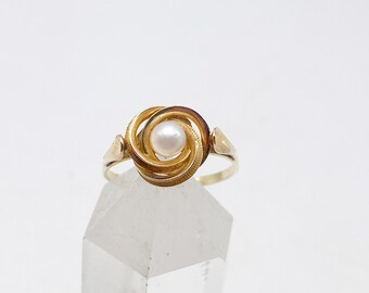 Op de afbeelding: Een gouden ring met een centrale witte parel omringd door een wervelend, gelaagd goudkleurig metalen ontwerp. De ringband is dun en gepolijst en rust op een heldere, puntige kristalstructuur tegen een effen witte achtergrond.
