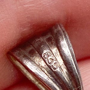 May include: Close-up of a silver pendant with a textured, fan-like design. The pendant has a small, stamped marking that reads "835". The metal has a slightly tarnished appearance.