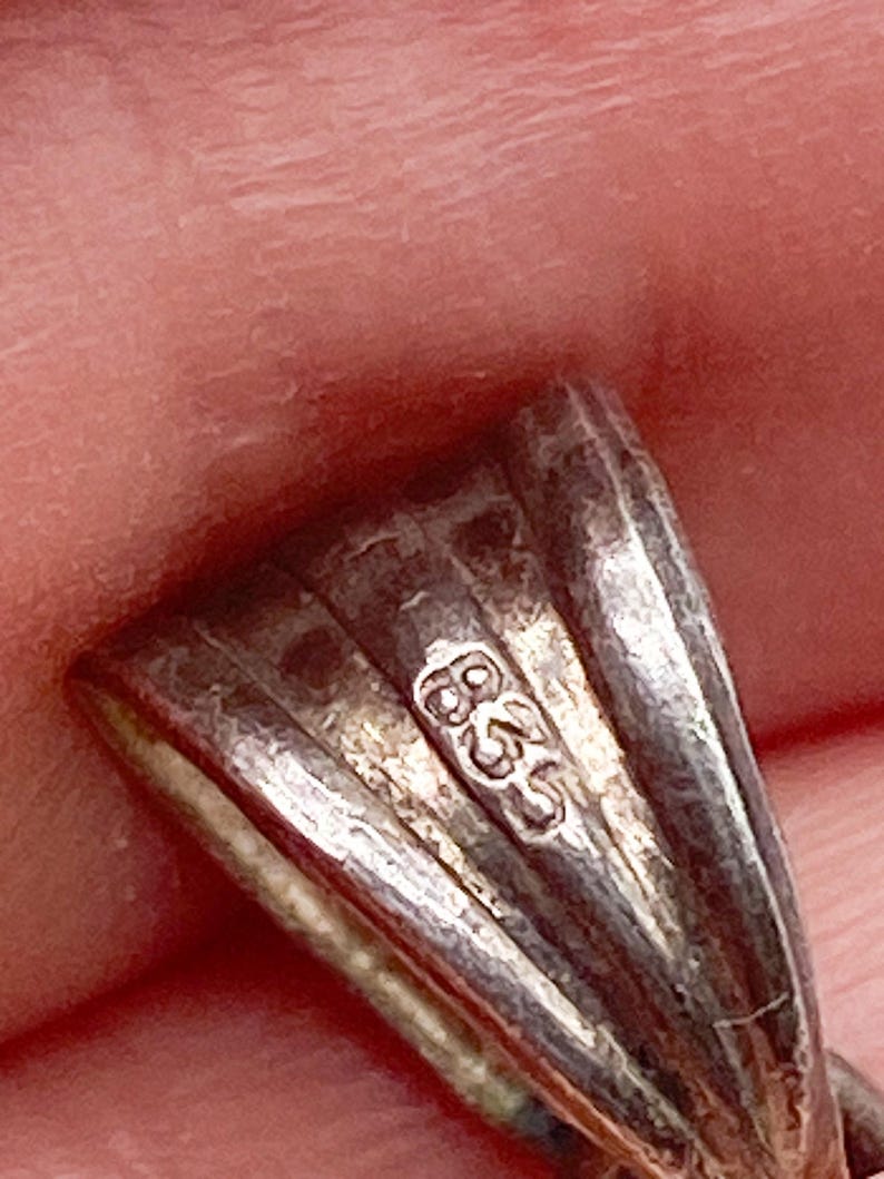 May include: Close-up of a silver pendant with a textured, fan-like design. The pendant has a small, stamped marking that reads "835". The metal has a slightly tarnished appearance.
