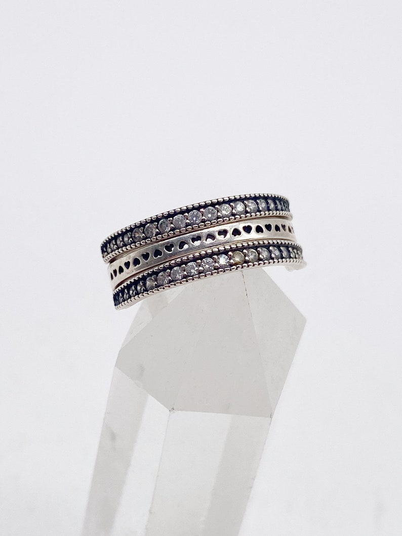 May include: A silver ring with three bands, each featuring a row of small clear stones and a delicate heart design.
