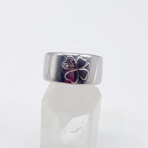 May include: A silver-toned ring with a wide band. The ring features a four-leaf clover design on the front, with small, clear stones set in the center of the clover. The ring is displayed on a clear, faceted surface.