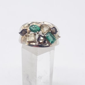 May include: A silver ring featuring an array of colorful gemstones. The ring showcases emerald green, clear, and dark gray stones in various shapes, including emerald cuts and teardrops. The ring is set on a clear, rectangular display stand against a white background.
