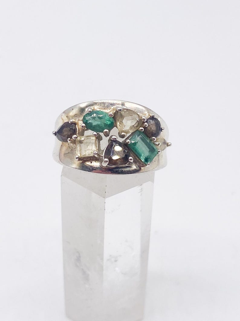 May include: A silver ring featuring an array of colorful gemstones. The ring showcases emerald green, clear, and dark gray stones in various shapes, including emerald cuts and teardrops. The ring is set on a clear, rectangular display stand against a white background.