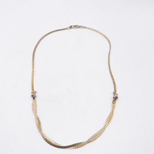 Può includere: Una collana a catena color oro caratterizzata da un design a spina di pesce intrecciato e piatto. La collana presenta due inserti metallici decorativi su entrambi i lati, ciascuno incastonato con piccole pietre multicolori, e una chiusura a moschettone.