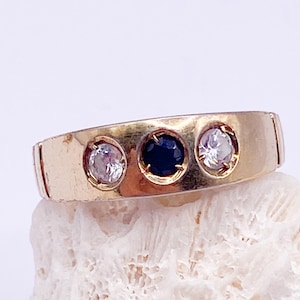 May include: A gold ring with three gemstones: a dark blue sapphire in the center, flanked by two clear stones. The ring has a simple band design and is set on a textured, off-white surface.