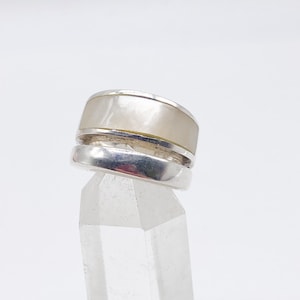Pode incluir: Um anel de prata com uma banda larga e plana e uma inserção de cor mais clara. O anel é exibido em um cristal claro e facetado. O design é simples e moderno, adequado para uso diário ou como peça de destaque.
