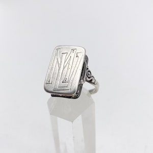 May include: A silver ring with a rectangular face featuring a stylized letter 'M' design. The ring has a delicate, ornate band.