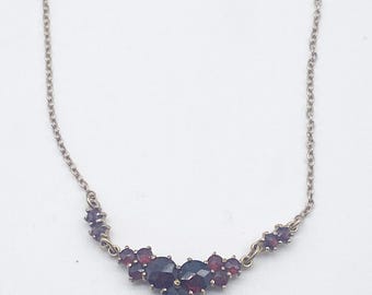 May include: A silver-toned necklace featuring a cluster of dark red gemstones. The gemstones are arranged in a curved design, with a larger central cluster and smaller stones extending to the sides. The chain is delicate and the necklace is set against a white background.