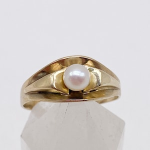 Könnte beinhalten: Ein goldener Ring mit einem einzelnen weißen Perlmutt, der in der Mitte eingesetzt ist. Der Ring hat ein einzigartiges, geschwungenes Design.