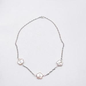 Pode incluir: Um colar prateado com três pérolas irregulares esbranquiçadas. As pérolas são conectadas por uma corrente delicada, criando um design simples, mas elegante. O colar é exibido em um fundo branco liso.