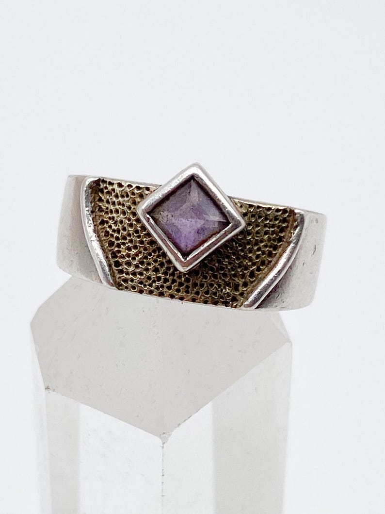May include: A silver ring featuring a textured gold band and a square, purple gemstone set in a silver bezel. The ring is displayed on a clear, faceted surface against a white backdrop.