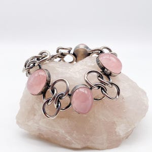 Puede incluir: Una pulsera de plata con piedras preciosas rosas ovaladas. La pulsera presenta un dise&ntilde;o de eslabones de cadena con las piedras preciosas engastadas en biseles individuales. La pulsera se exhibe sobre una piedra rosa p&aacute;lido de textura rugosa.