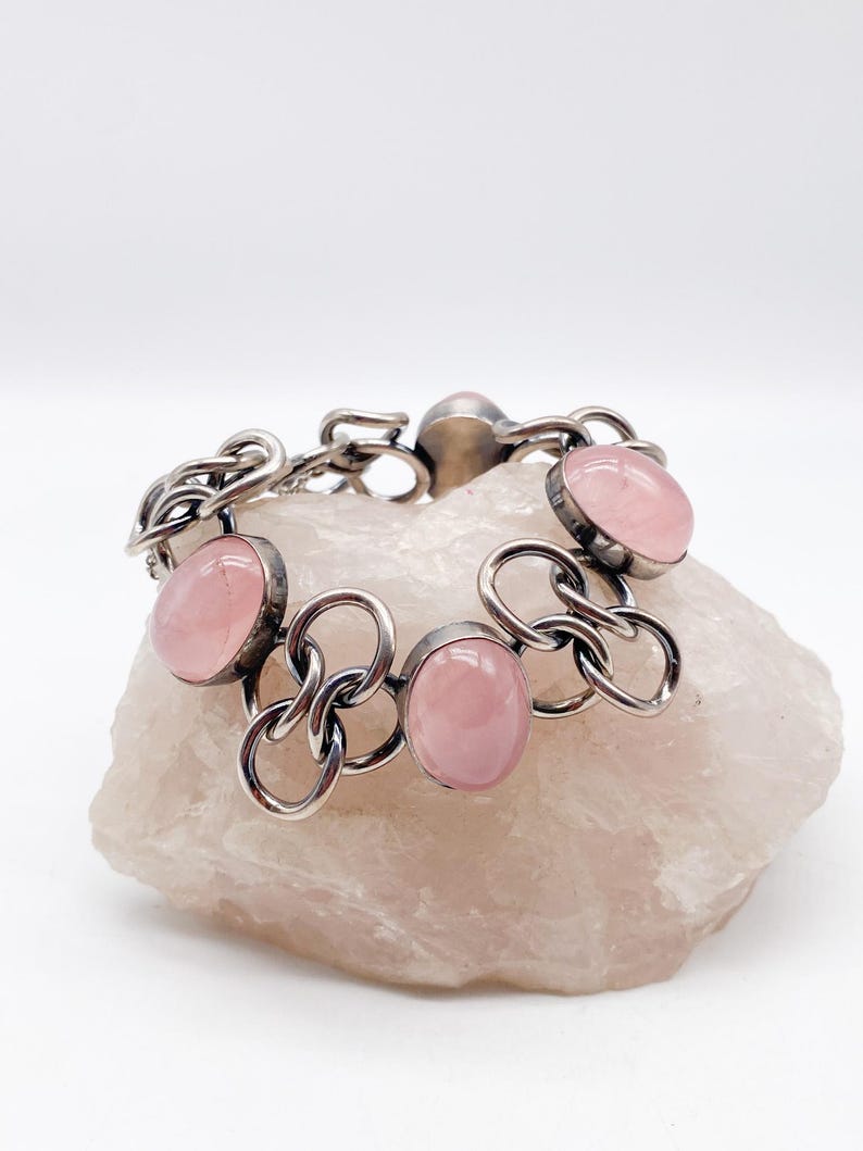 Puede incluir: Una pulsera de plata con piedras preciosas rosas ovaladas. La pulsera presenta un dise&ntilde;o de eslabones de cadena con las piedras preciosas engastadas en biseles individuales. La pulsera se exhibe sobre una piedra rosa p&aacute;lido de textura rugosa.