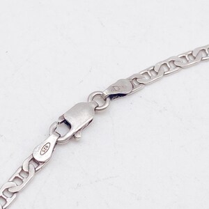 May include: Close-up of a silver chain necklace with a lobster clasp. The chain has a flat curb link design. The clasp is marked with "925". The necklace is displayed against a white background.