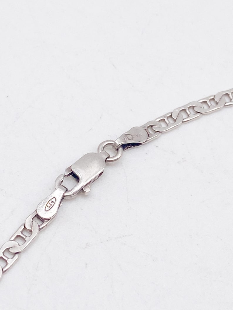 May include: Close-up of a silver chain necklace with a lobster clasp. The chain has a flat curb link design. The clasp is marked with "925". The necklace is displayed against a white background.