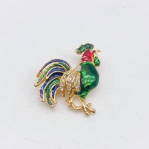 Può includere: Spilla a forma di gallo colorato con base in metallo dorato. Il gallo presenta dettagli in smalto verde, blu e rosso, con accenti di cristallo. Un accessorio decorativo.
