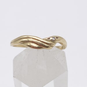 Puede incluir: Un anillo de oro con un diseño fluido e entrelazado. El anillo presenta una pequeña gema transparente engastada en la parte superior del diseño. El anillo se muestra sobre una superficie clara y facetada sobre un fondo blanco.