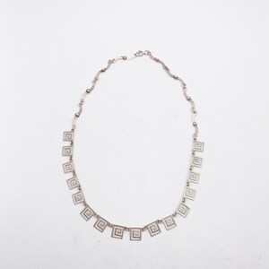 May include: A silver necklace with a chain made of small, linked squares. The necklace has a series of larger, square pendants with a geometric design.