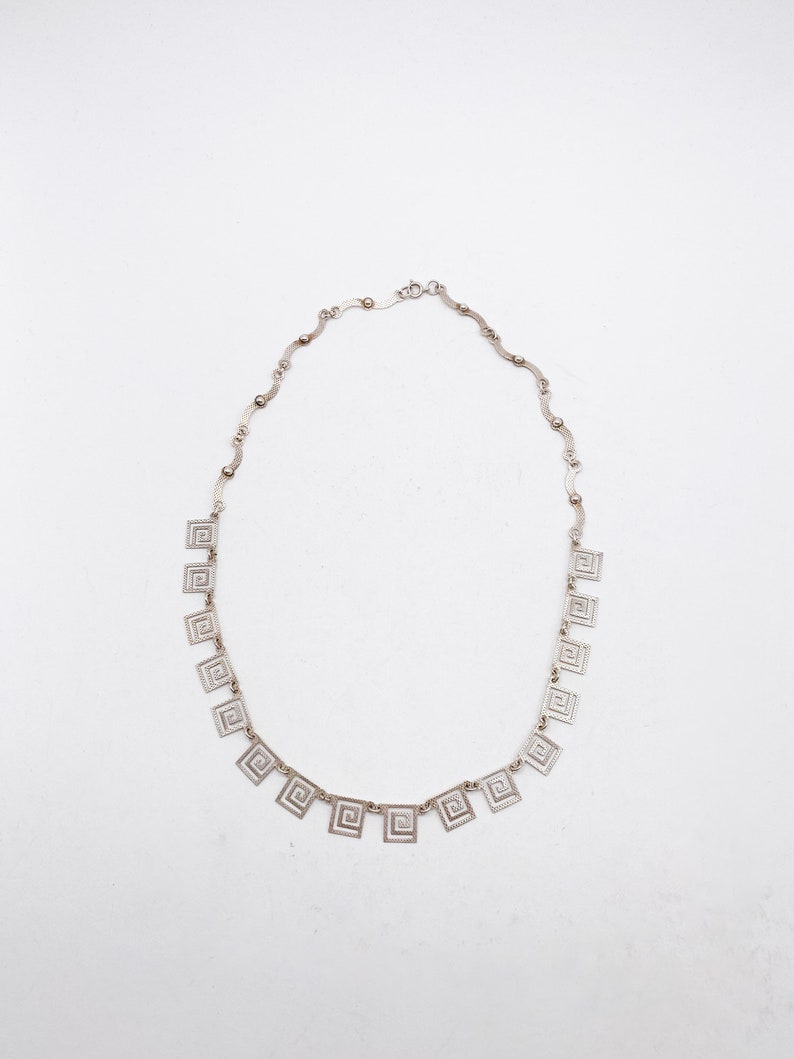 May include: A silver necklace with a chain made of small, linked squares. The necklace has a series of larger, square pendants with a geometric design.