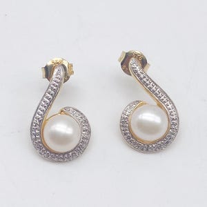 May include: A pair of gold-toned drop earrings with a white pearl center and a swirl design encrusted with small, clear stones. The earrings have a post-back closure.