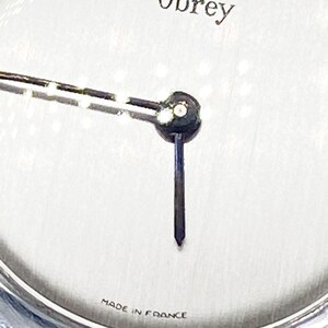 May include: A silver watch face with black hands and the word "Obrey" printed in black at the top. The watch face also has the words "Made in France" printed at the bottom.