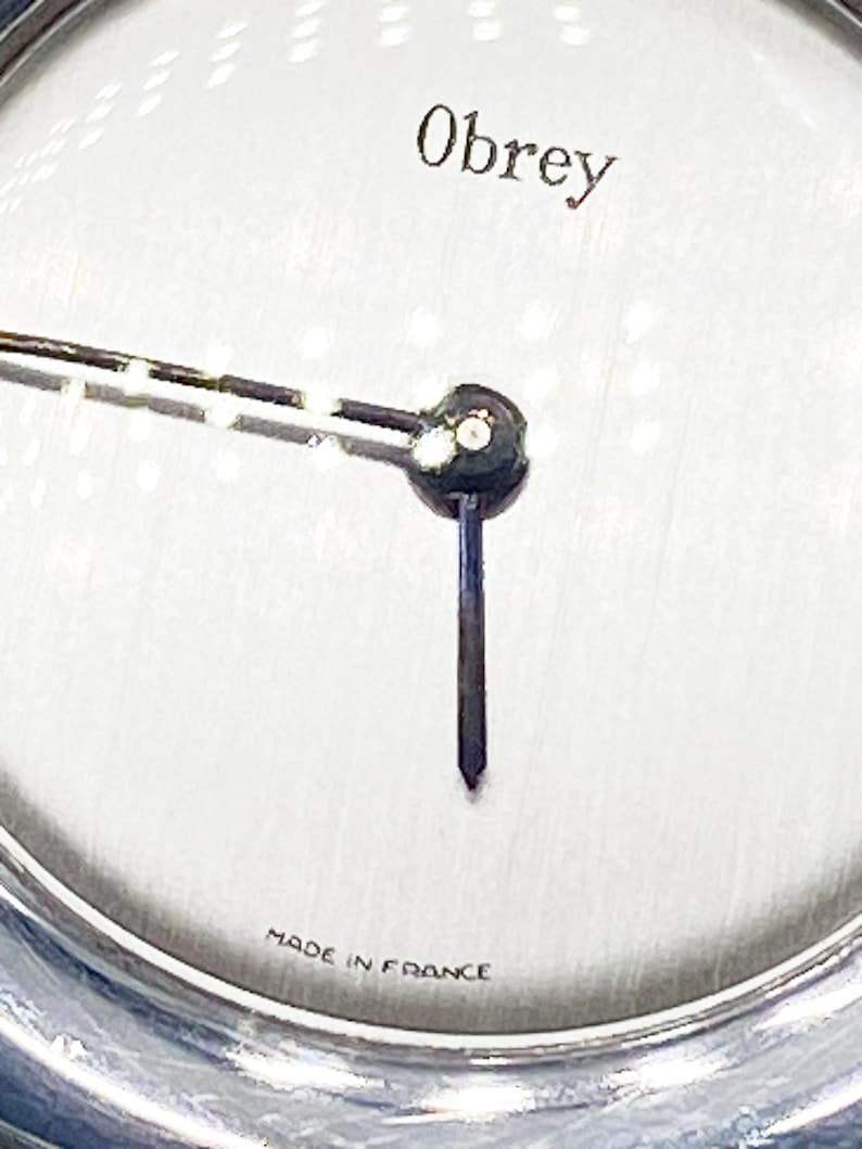 May include: A silver watch face with black hands and the word "Obrey" printed in black at the top. The watch face also has the words "Made in France" printed at the bottom.