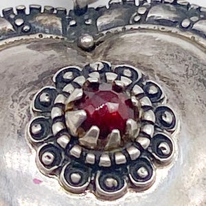 May include: Close-up of a silver heart-shaped pendant with a decorative border. The pendant features a central, round, deep red gemstone surrounded by a detailed floral design. The metal has an aged, antique appearance.