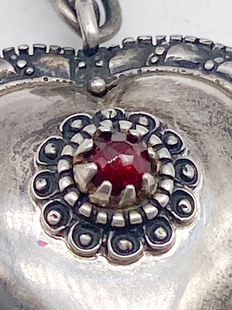 May include: Close-up of a silver heart-shaped pendant with a decorative border. The pendant features a central, round, deep red gemstone surrounded by a detailed floral design. The metal has an aged, antique appearance.