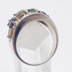 May include: A silver ring with a row of rectangular gemstones in shades of green, blue, and yellow. The ring has a wide band and a polished finish. The gemstones are set in a row across the top of the ring.