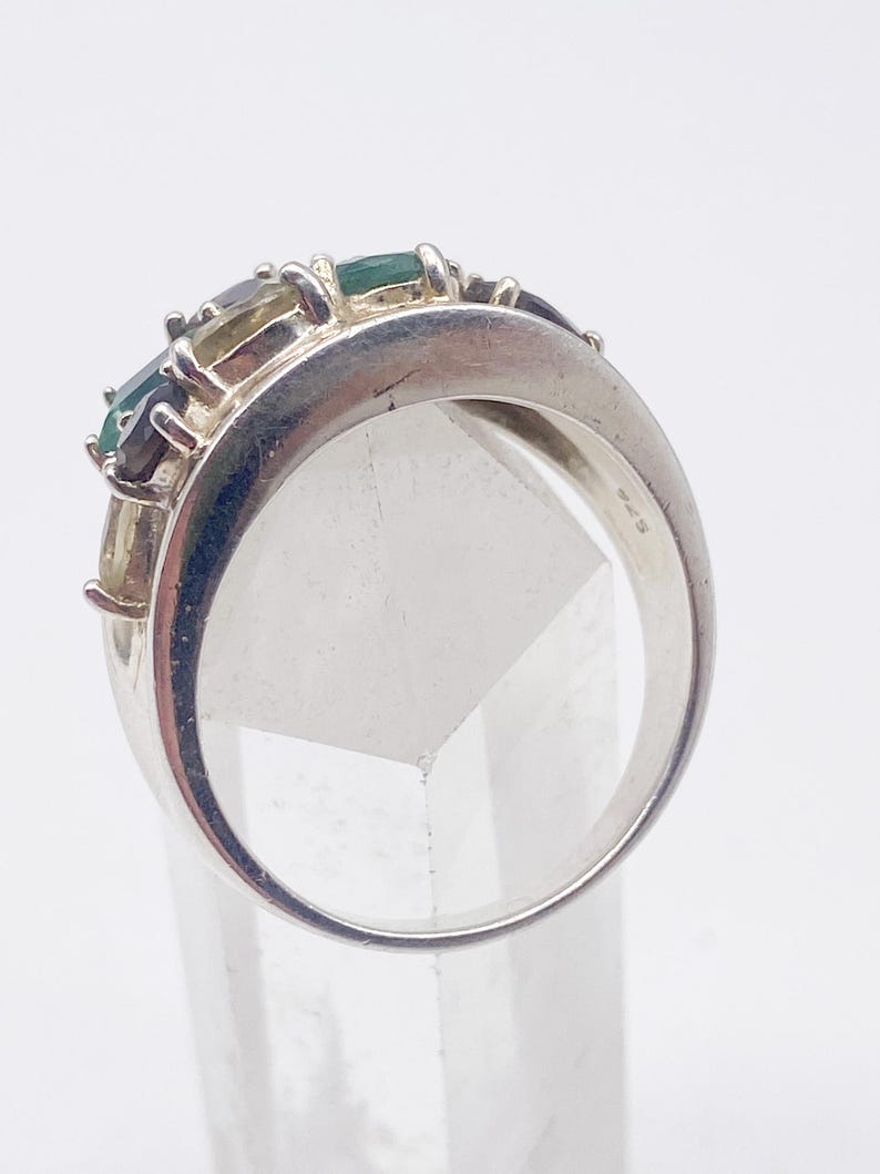 May include: A silver ring with a row of rectangular gemstones in shades of green, blue, and yellow. The ring has a wide band and a polished finish. The gemstones are set in a row across the top of the ring.