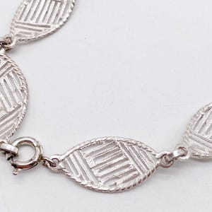 May include: A silver bracelet with a delicate, patterned design. The bracelet is made of oval-shaped links that are connected by small, round links. The bracelet has a clasp closure.