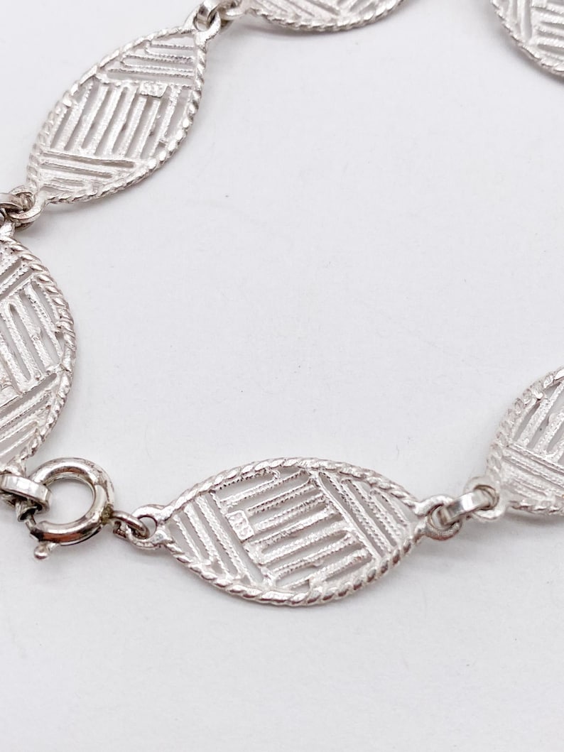 May include: A silver bracelet with a delicate, patterned design. The bracelet is made of oval-shaped links that are connected by small, round links. The bracelet has a clasp closure.