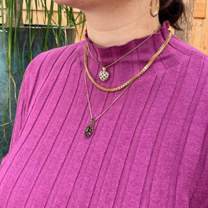 Pu&ograve; includere: Una donna indossa un maglione a collo alto a costine viola con tre collane d'oro. Le collane presentano un piccolo ciondolo a forma di cuore con pietre chiare, un ciondolo a forma di lacrima con pietre nere e un design a catena.