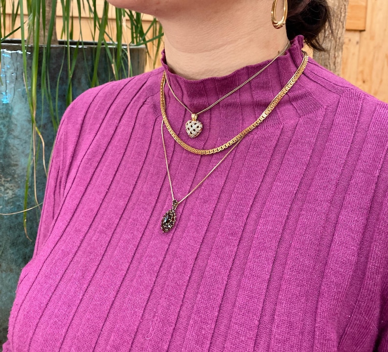 Pu&ograve; includere: Una donna indossa un maglione a collo alto a costine viola con tre collane d'oro. Le collane presentano un piccolo ciondolo a forma di cuore con pietre chiare, un ciondolo a forma di lacrima con pietre nere e un design a catena.