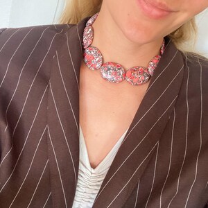 May include: A statement necklace featuring smooth, oval-shaped beads with a mottled pattern of red, black, and white. The necklace is worn with a dark brown blazer featuring thin white pinstripes and a white top with a gathered, ruched neckline.