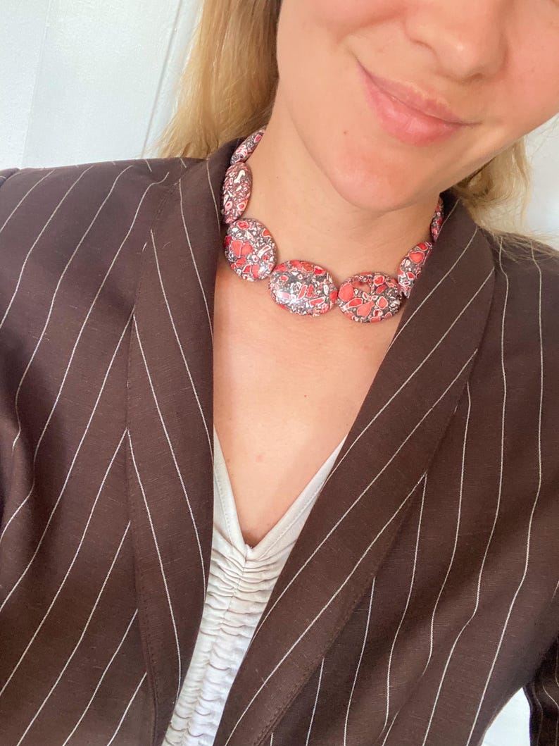 May include: A statement necklace featuring smooth, oval-shaped beads with a mottled pattern of red, black, and white. The necklace is worn with a dark brown blazer featuring thin white pinstripes and a white top with a gathered, ruched neckline.