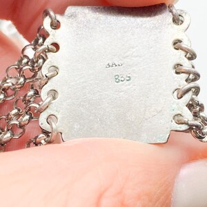 May include: Close-up of a silver metal jewelry piece with chain links. The piece has a rectangular plate with the markings "BAD" and "835" stamped on it. The chain links are attached to the plate with small loops.