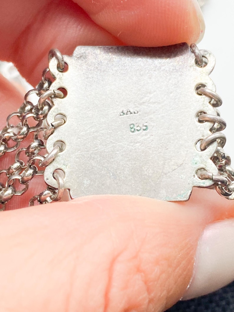 May include: Close-up of a silver metal jewelry piece with chain links. The piece has a rectangular plate with the markings "BAD" and "835" stamped on it. The chain links are attached to the plate with small loops.